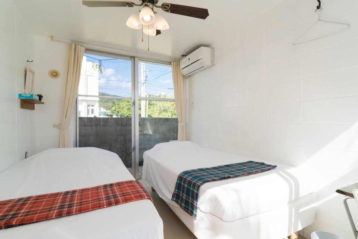 Ein Hotelzimmer mit zwei Betten, einem Deckenventilator, einer Klimaanlage, einem Fenster mit Vorhängen und Blick auf Gebäude.