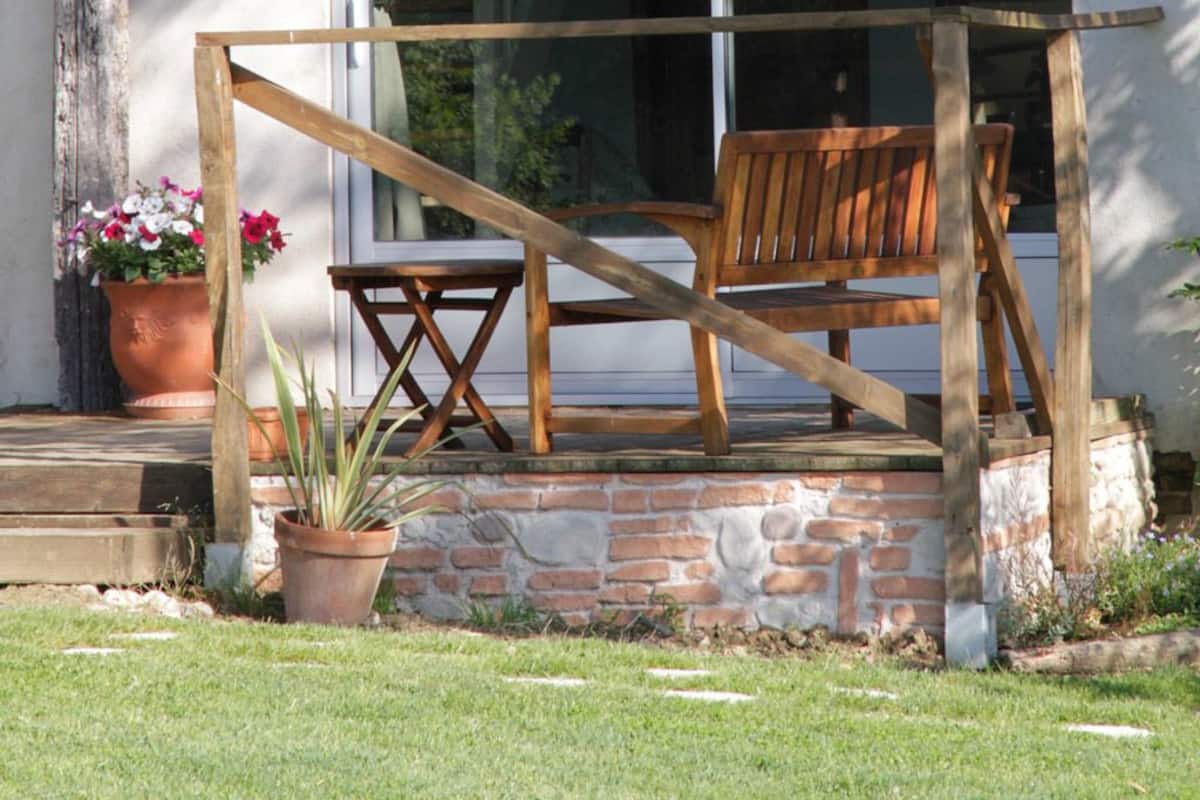 A wooden bench and chair on a porch with a stone wall and potted plants.