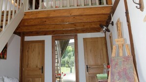 Una habitación con escalera de madera, un altillo, un cuadro sobre caballete y vistas al exterior.