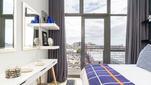 Ett hotellrum med ett stort fönster, en säng med ett blå- och vitrutigt överkast, ett vitt skrivbord med en spegel och utsikt över stadsbilden.