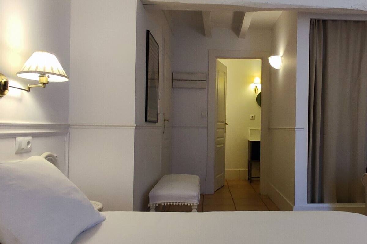 A hotel room with a bed, two armchairs, a small stool, a mirror, and a window with curtains.
