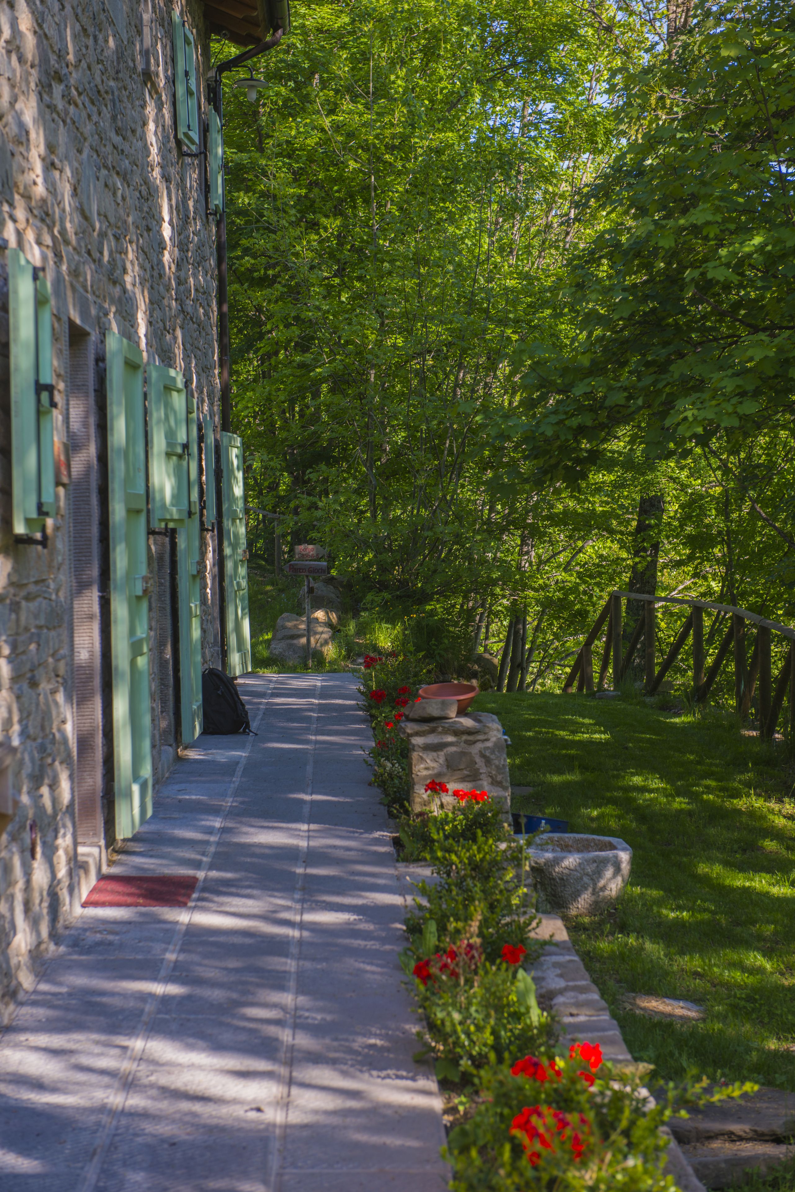 A stone building with green shutters, a paved pathway, and a garden with red flowers.
