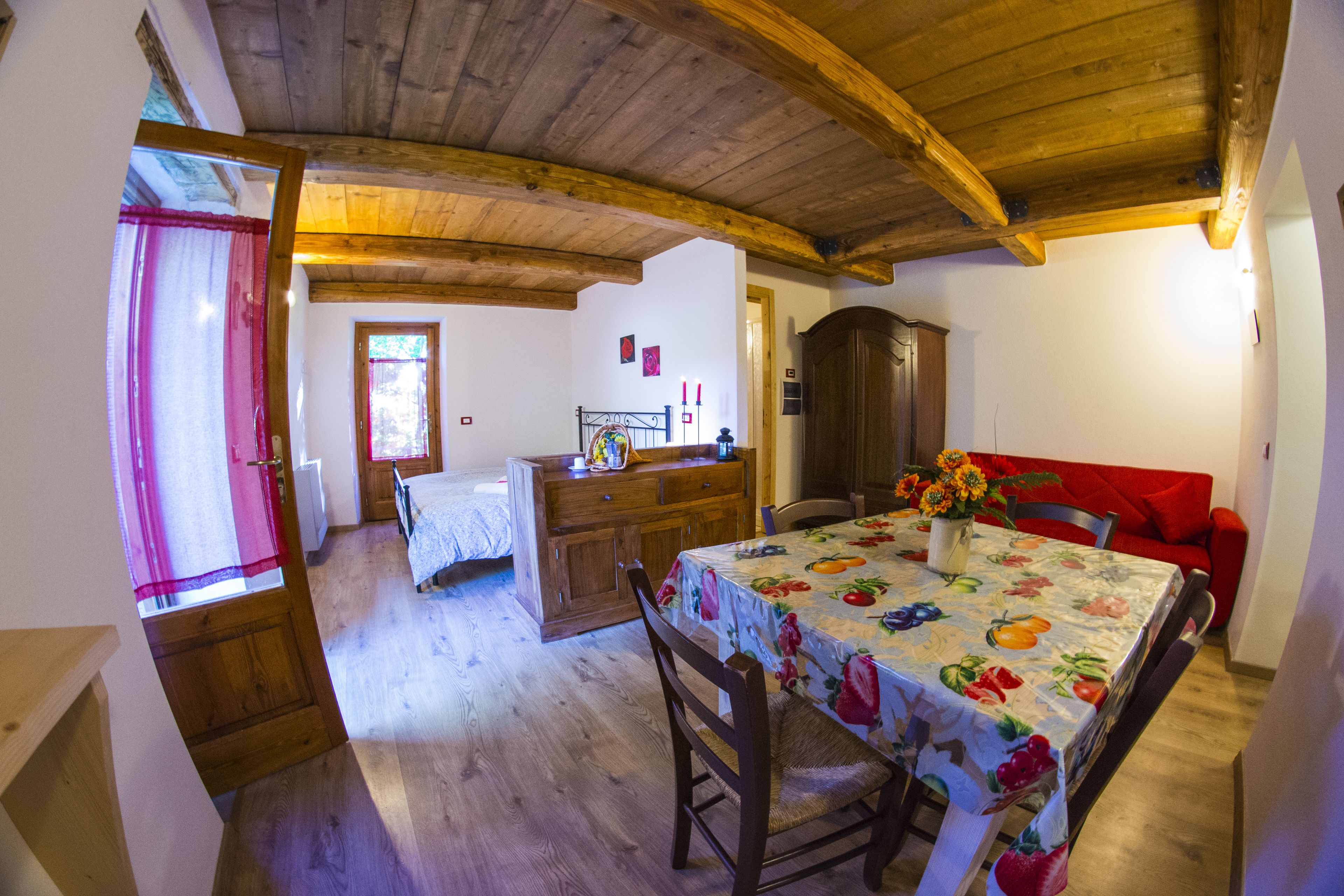 A cozy room with wooden beams, a dining table with a fruit pattern tablecloth, a red sofa, and a wooden cabinet.