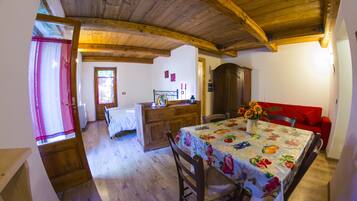 A cozy room with wooden beams, a dining table with a fruit pattern tablecloth, a red sofa, and a wooden cabinet.