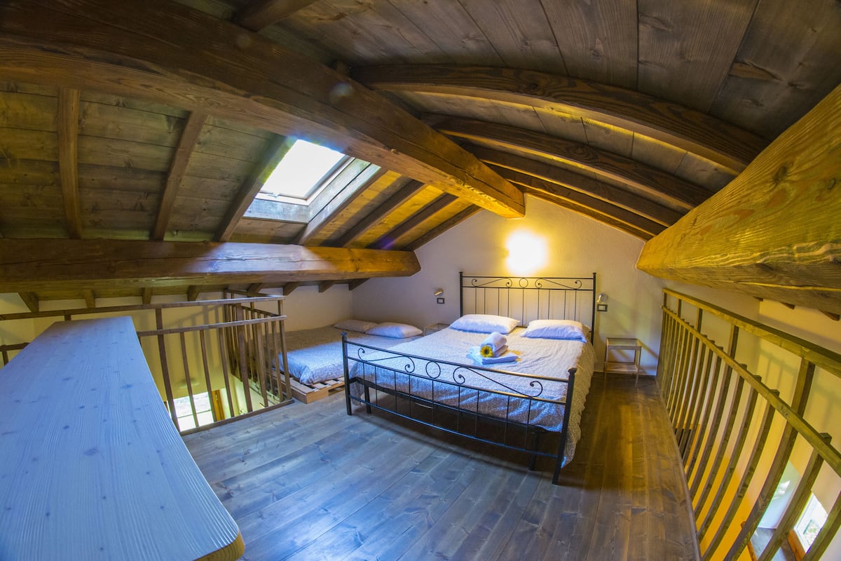 Une chambre à coucher aménagée dans un espace de type « loft », avec un plafond en bois, une lucarne, un lit avec des oreillers et un banc en bois.