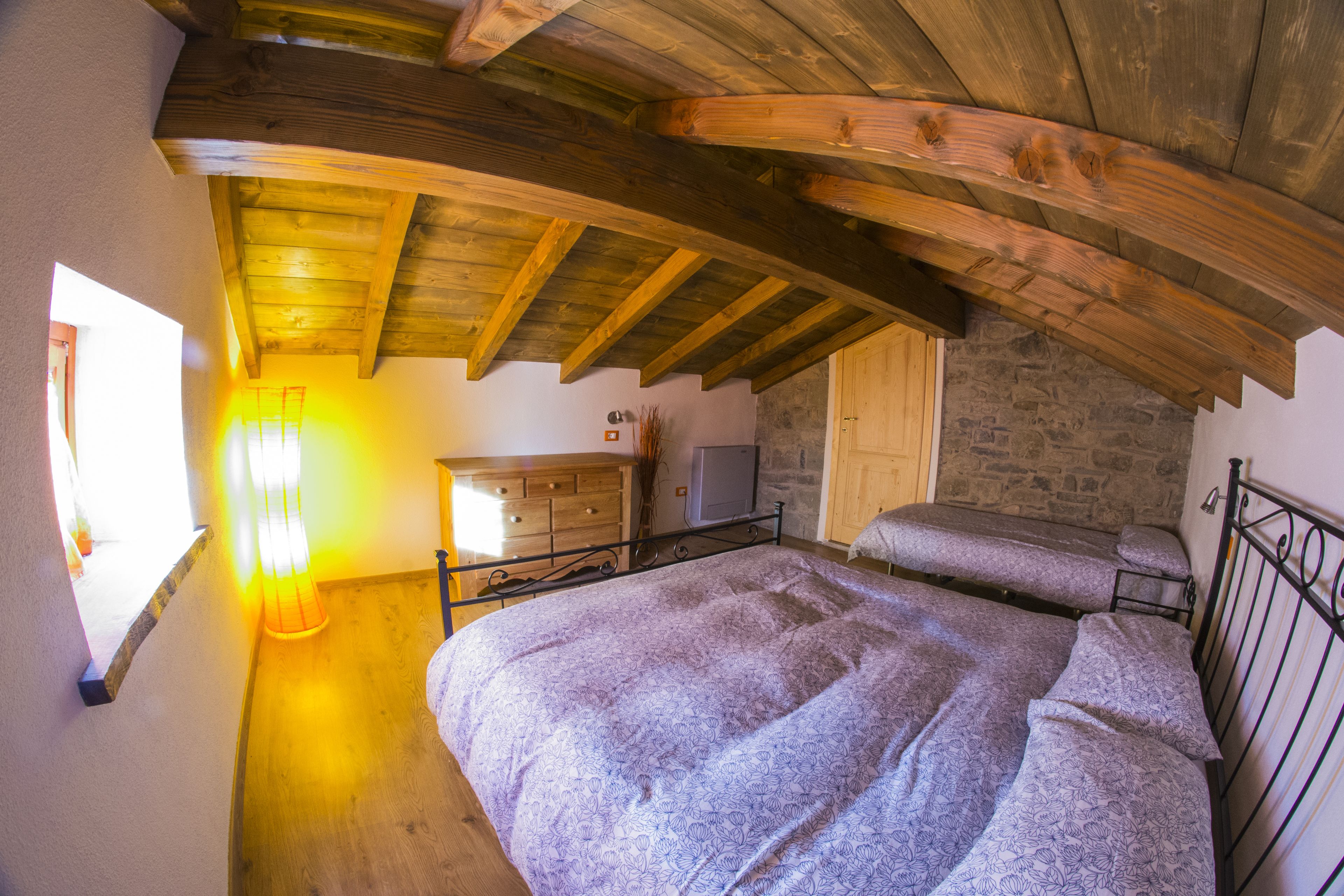 A bedroom with a wooden ceiling, a bed with patterned bedding, a dresser, and a stone wall.