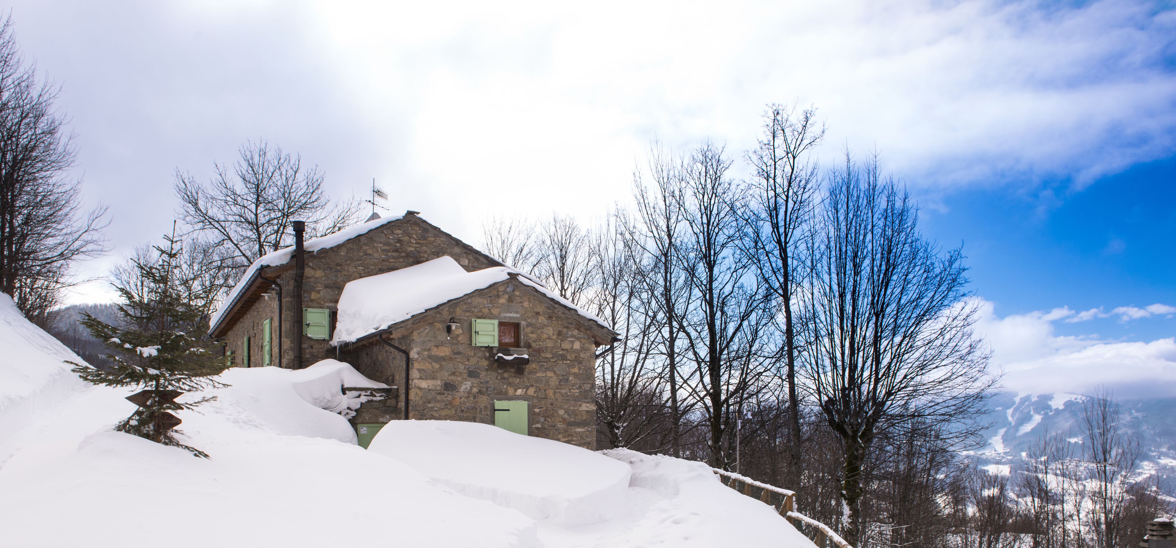 Ein Steinhaus mit schneebedecktem Dach und Fenstern, umgeben von kahl stehenden Bäumen und einer verschneiten Landschaft.
