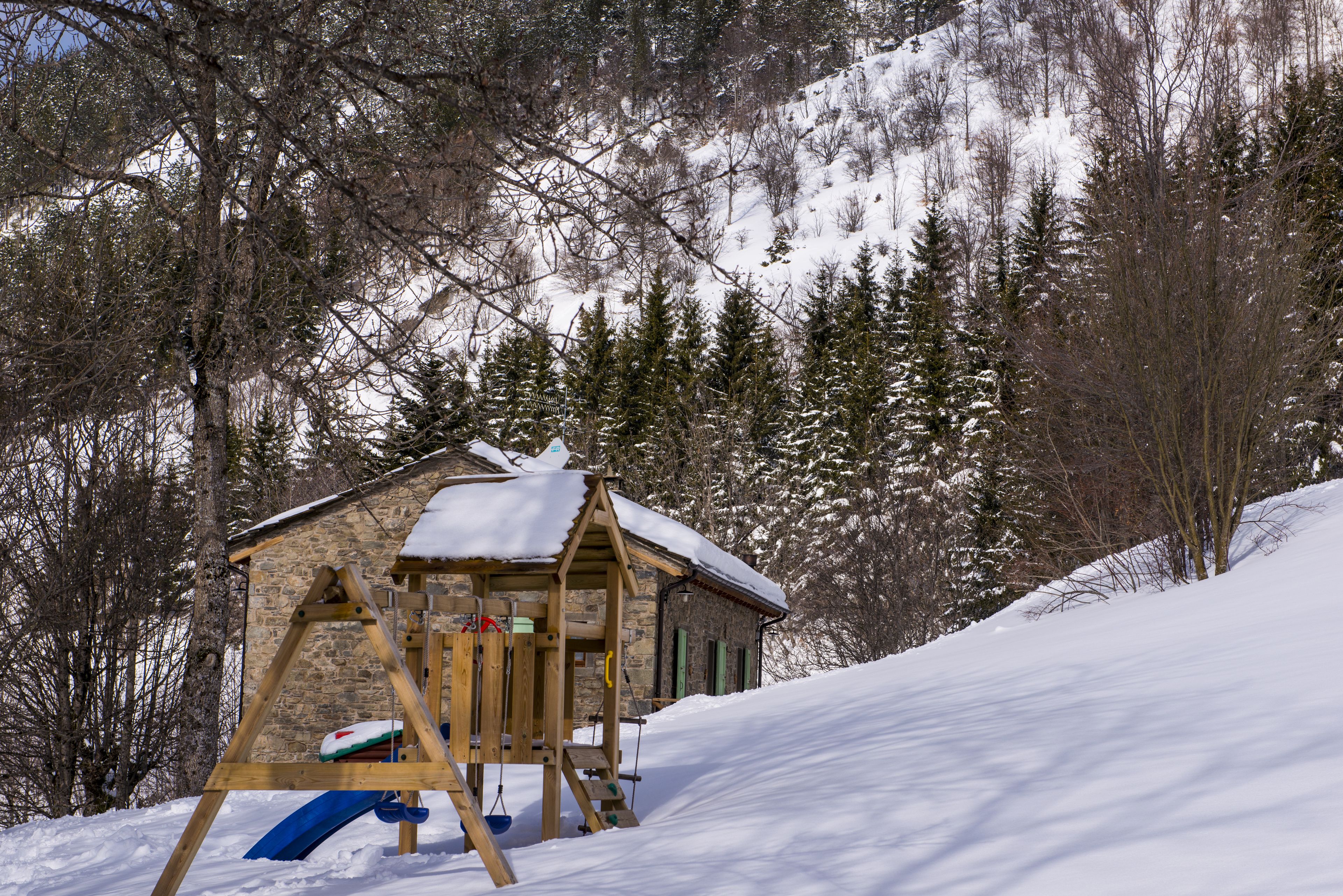 A wooden play structure with a slide, set against a snowy backdrop with trees and a stone building.
