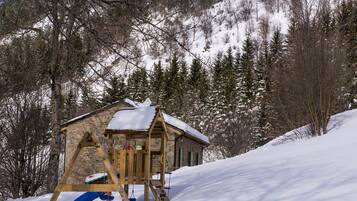 A wooden play structure with a slide, set against a snowy backdrop with trees and a stone building.
