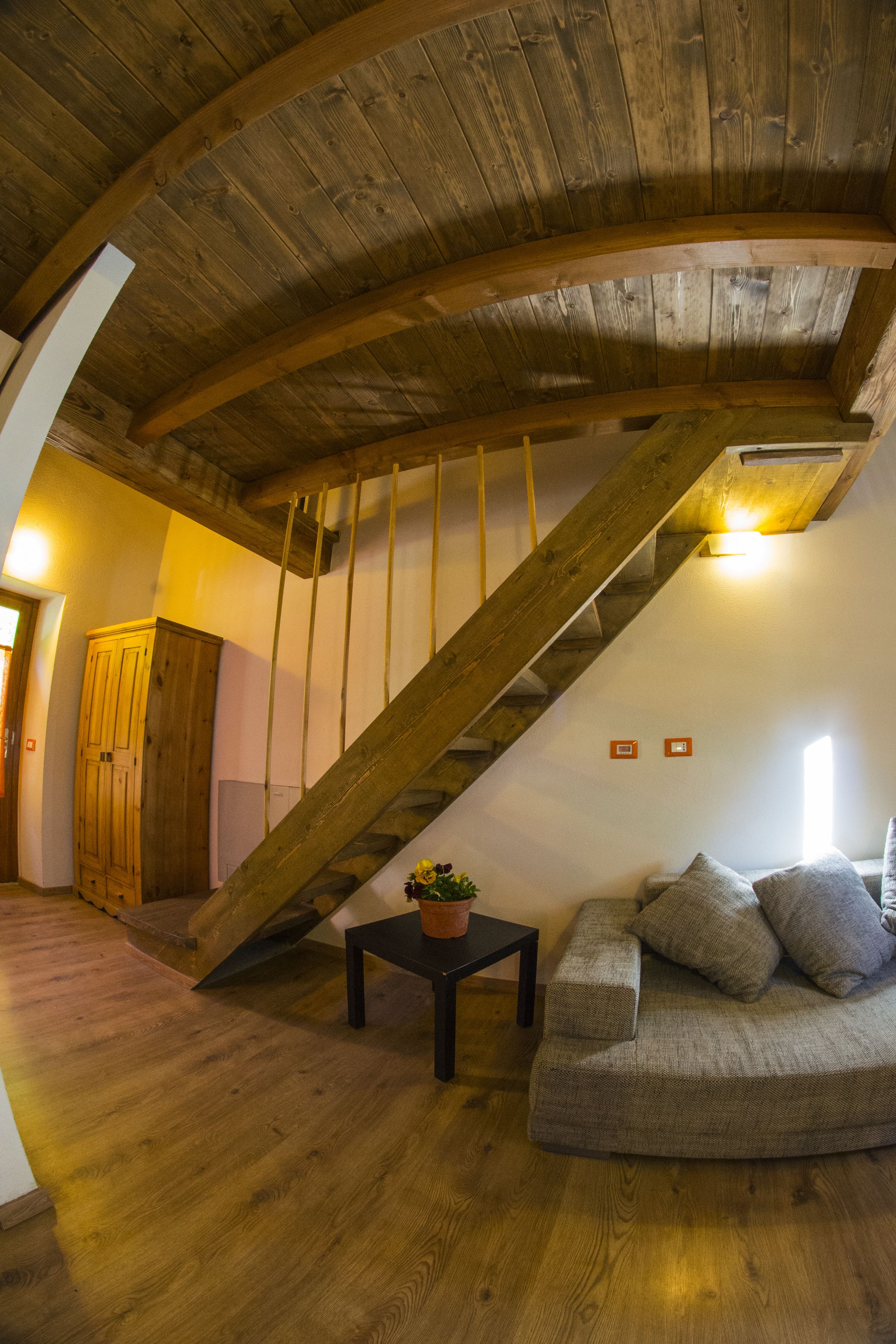 A wooden staircase with a curved design, a wooden floor, a sofa with cushions, and a small table with a flower vase.