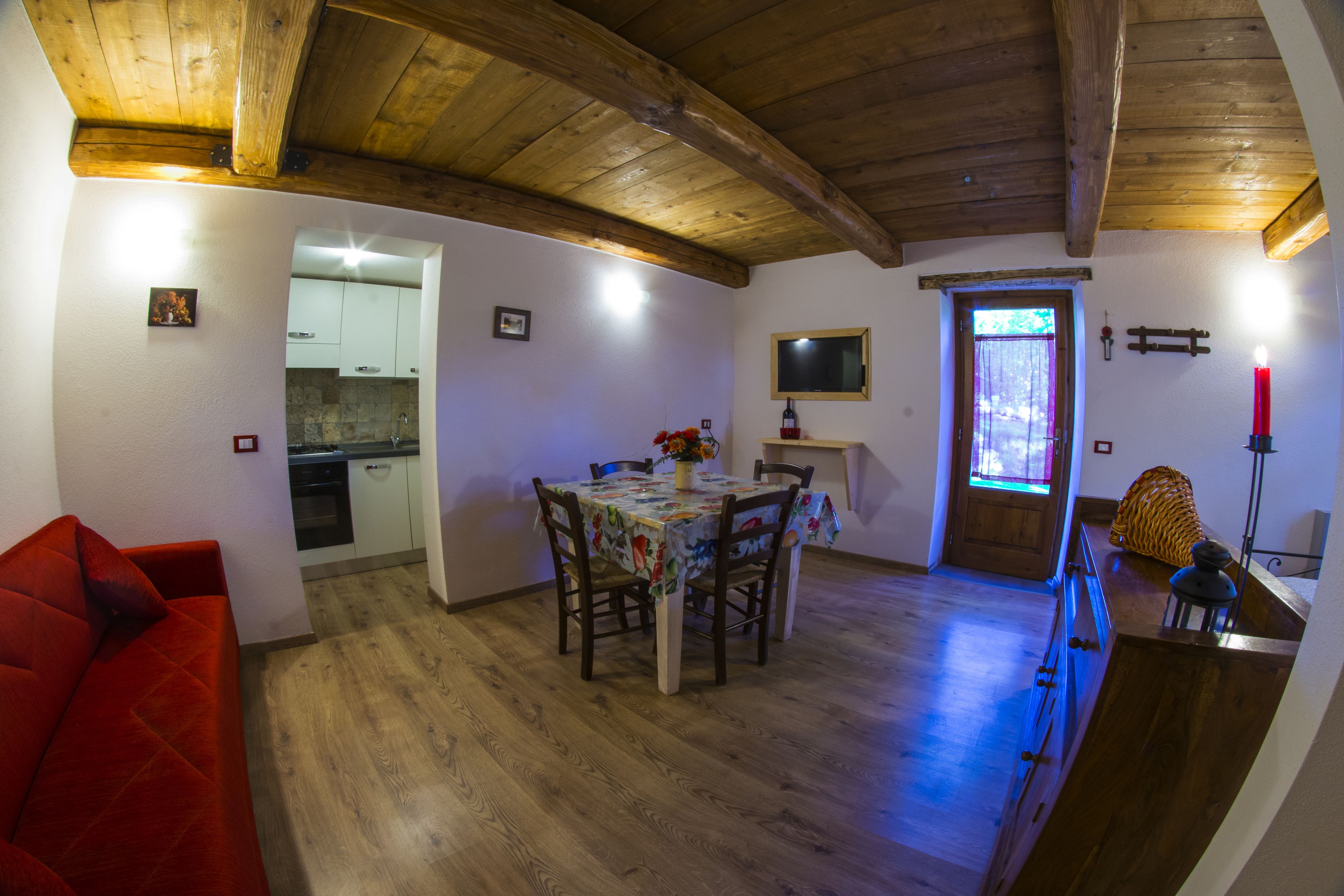 A cozy living room with a red sofa, a wooden dining table with chairs, and a kitchen in the background.