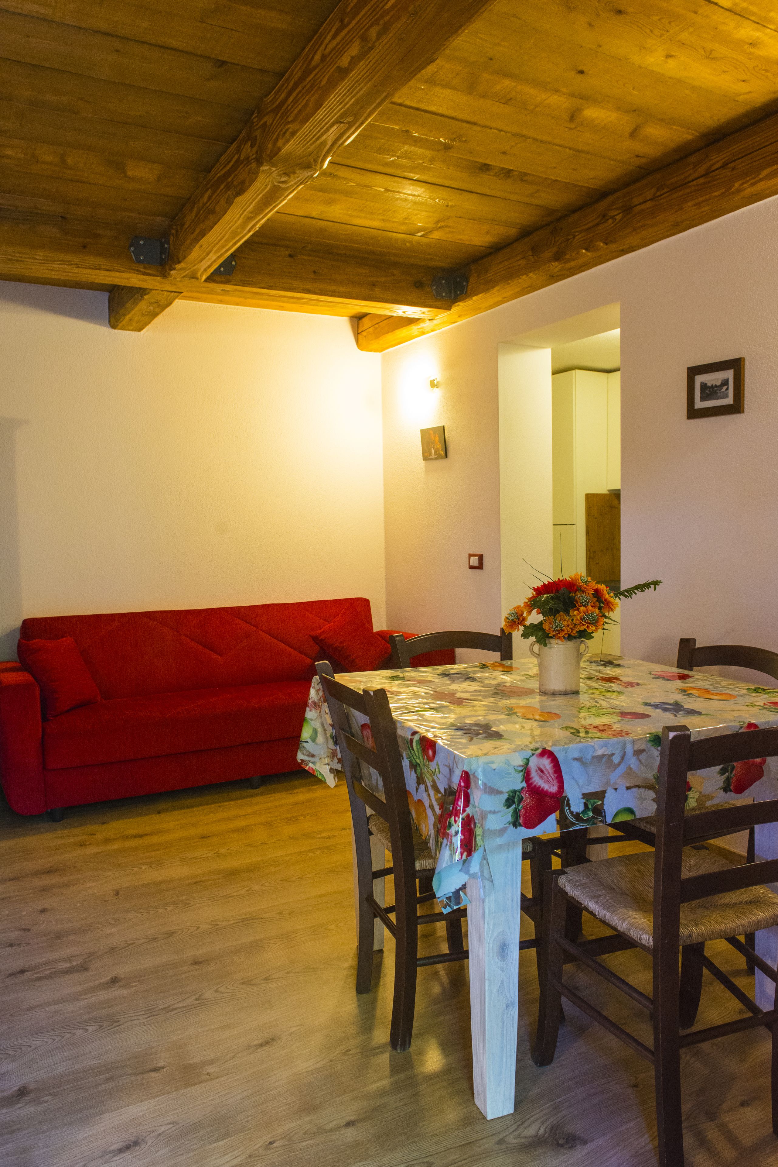 A dining area with a wooden ceiling, a red sofa, a table with a floral tablecloth, and a vase with flowers.
