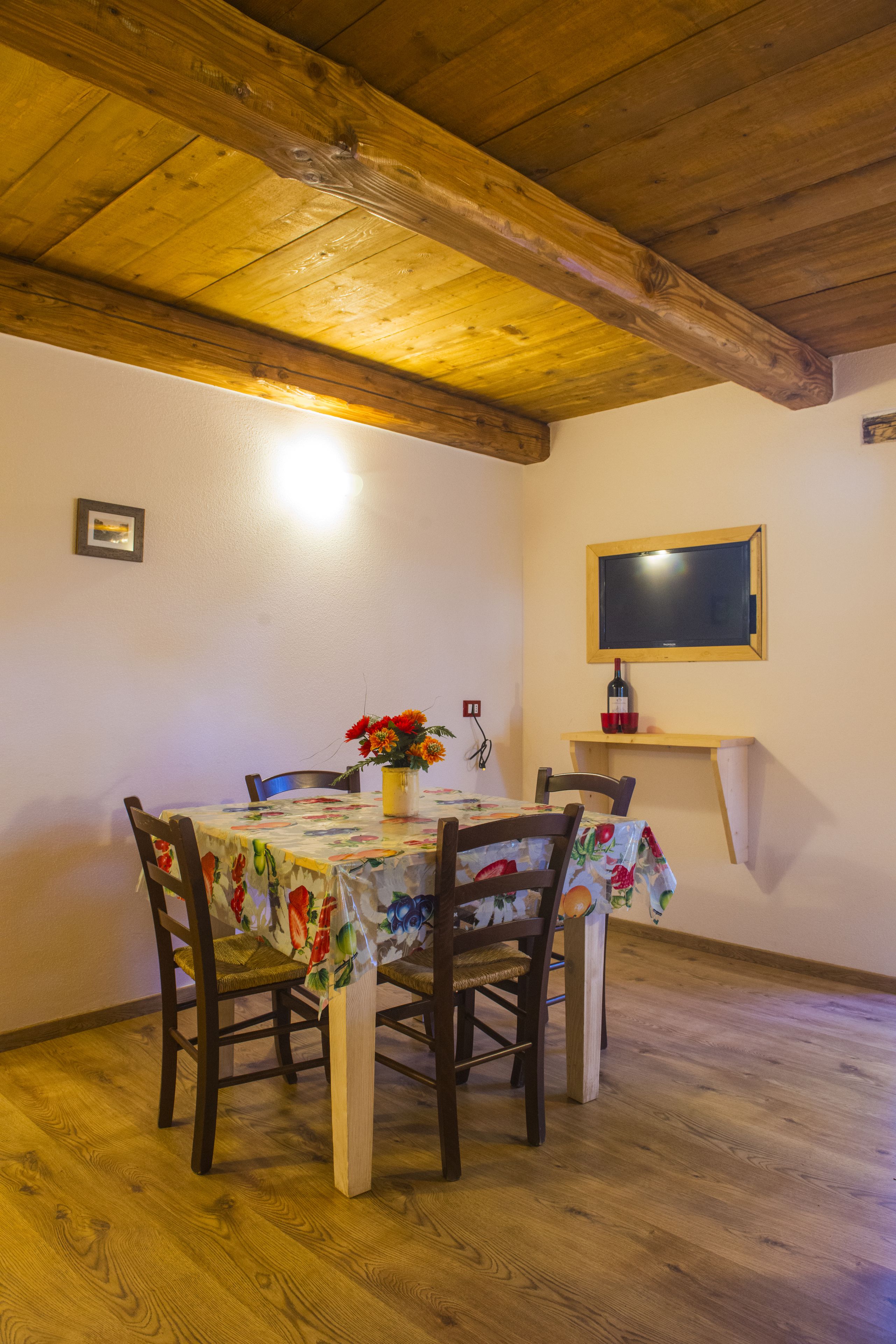 A dining area with a wooden ceiling, a table with a floral tablecloth, chairs, a vase with flowers, and a wall-mounted TV.