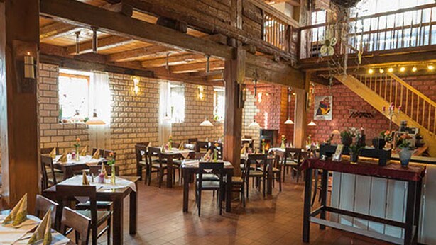 Um restaurante amplo com pé-direito alto, vigas de madeira aparentes, paredes de tijolo e uma escadaria que leva a um andar superior. As mesas estão dispostas com toalhas e guardanapos.