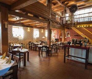 A spacious restaurant with high ceilings, exposed wooden beams, brick walls, and a staircase leading to an upper level. Tables are set with tablecloths and napkins. - Wellnesshotel und Restaurant Hexenschopf (Löffingen)