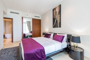 A modern hotel room with a large bed, a nightstand with a lamp, a bathroom with a bathtub, and a large mirror. - Deluxe Holiday Homes - Marina Gate (Dubai)