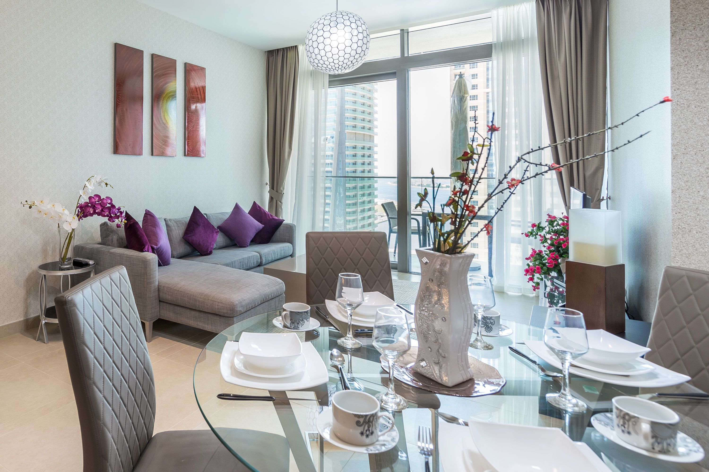 A modern living room with a glass dining table set for four, a grey sofa with purple cushions, and a city view through large windows.
