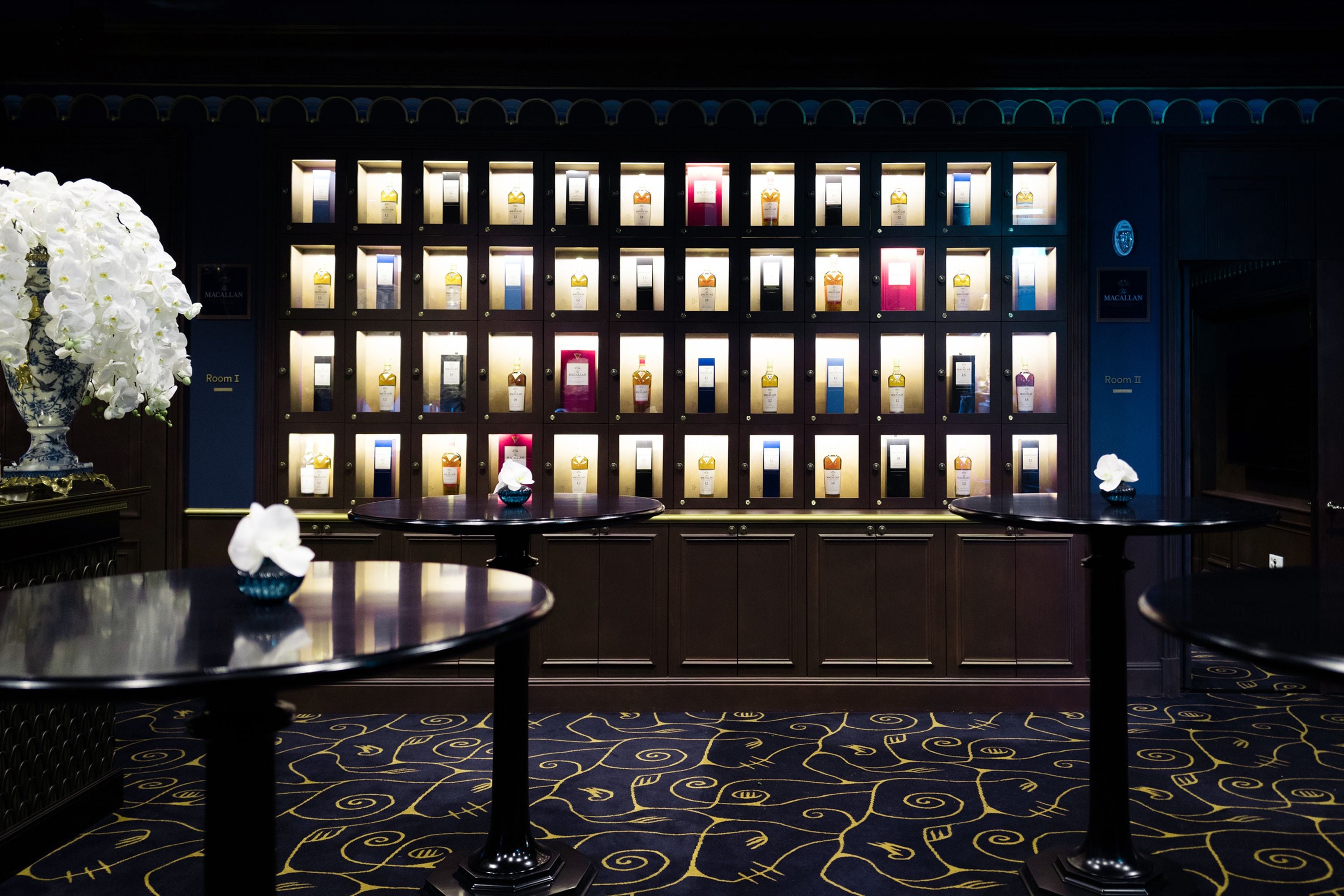 Un vestíbulo de hotel con una pared de vitrinas iluminadas, tres mesas redondas y una alfombra estampada.