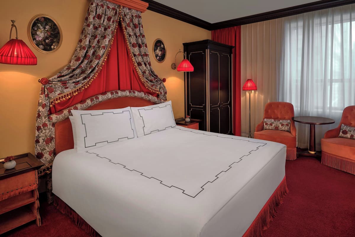 A hotel room with a large bed, a wardrobe, two armchairs, a small table, and a red carpet.