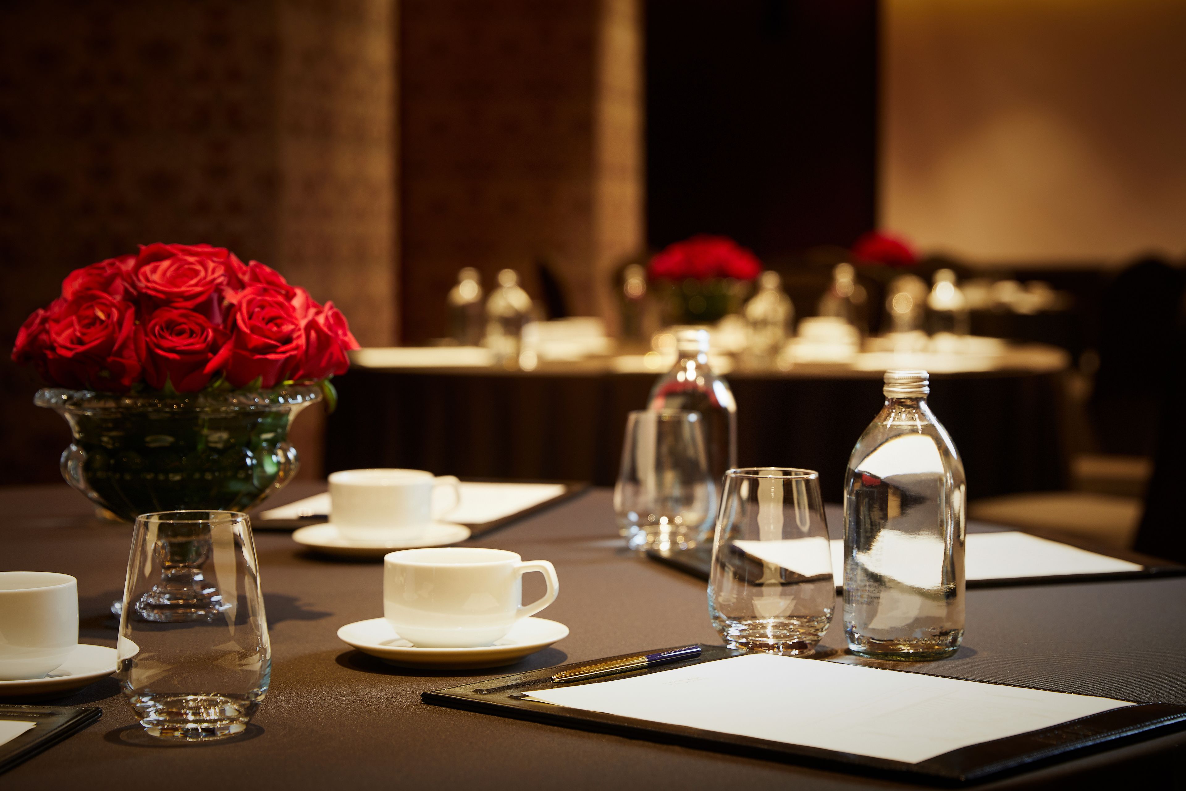 A table set for a meeting with a vase of red roses, clear glasses, and white cups.