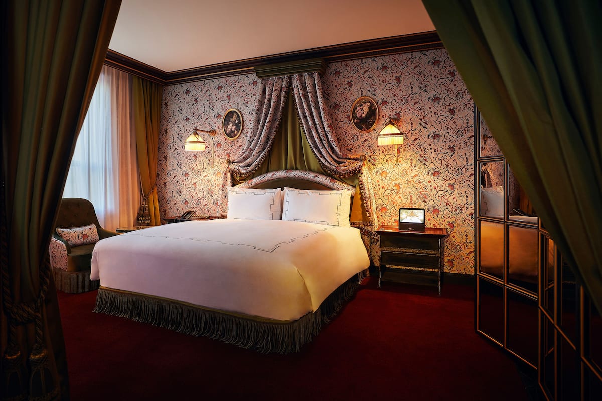 A large bed with a white bedspread and a dark fringe, flanked by two nightstands with lamps, in a room with patterned wallpaper and green curtains.