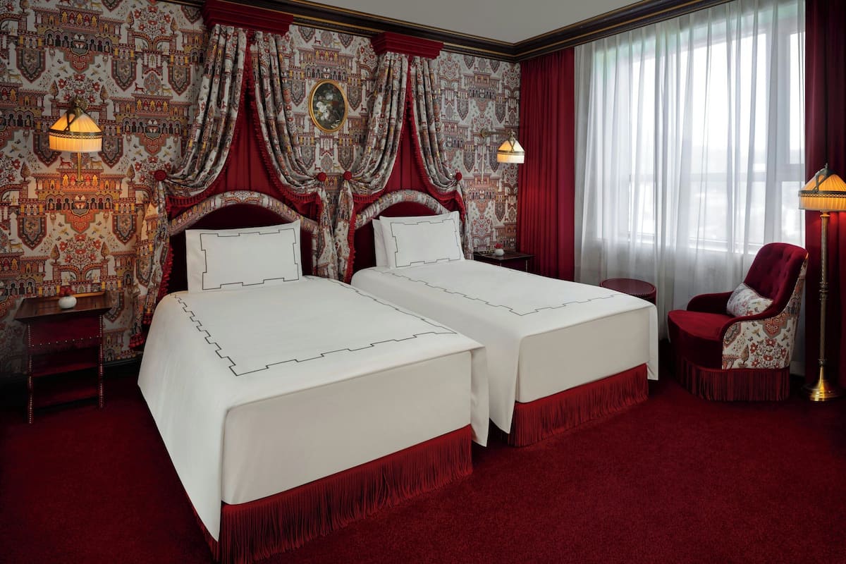 A hotel room with two beds, a red carpet, a red chair, and patterned wallpaper.