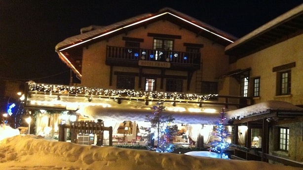 A hotel with a snow-covered roof, illuminated by string lights, and a balcony with railings. - Hôtel Franz (Allos)