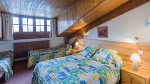 A bedroom with three beds, wooden ceiling beams, a window with curtains, and a bedside lamp. - Hôtel Franz (Allos)