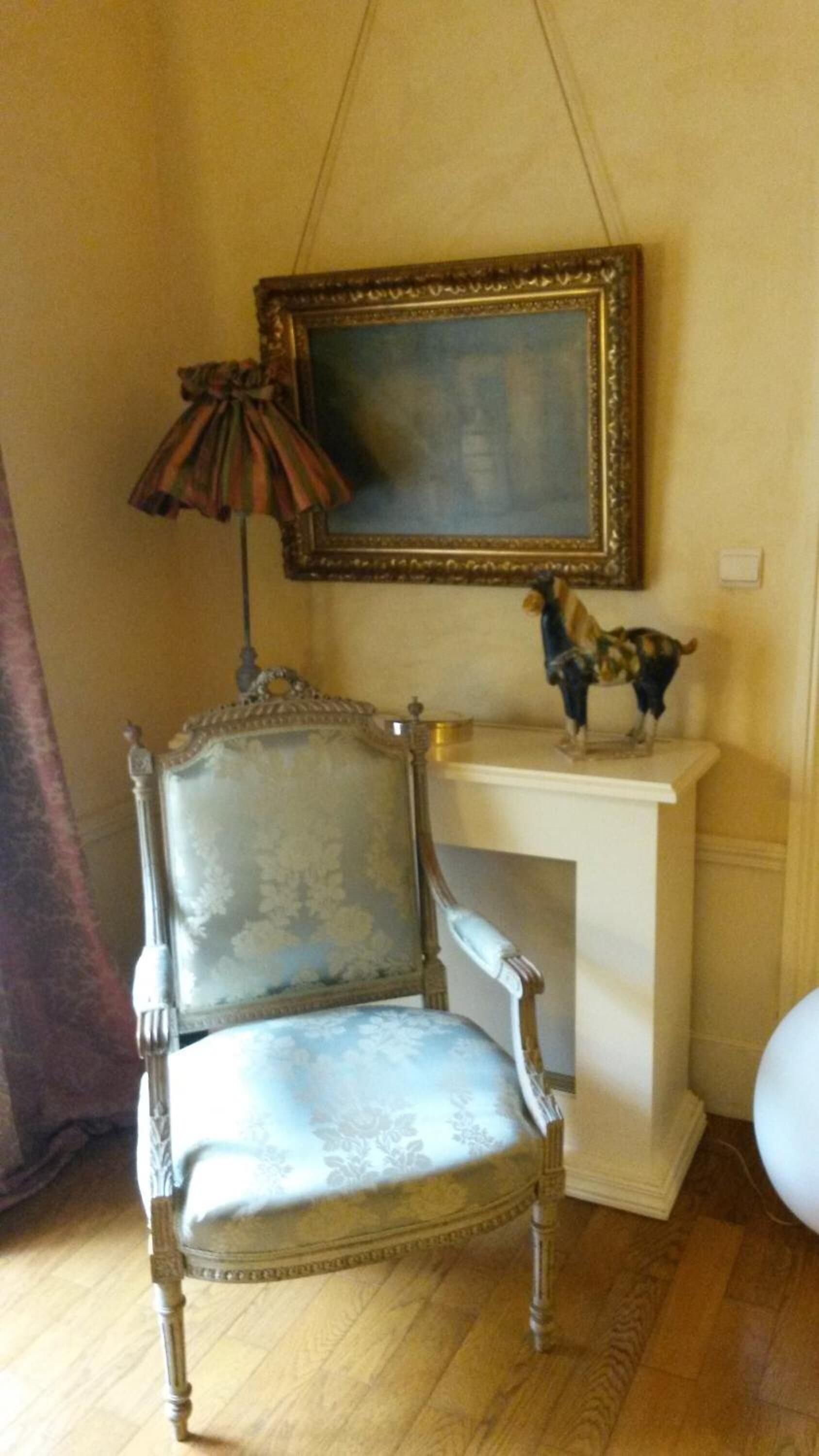 Een kamer met een houten vloer, een vintage stoel, een lamp, een ingelijst schilderij en een klein beeld op een witte sokkel.