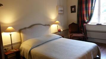 A neatly made bed with a headboard, two bedside lamps, a chair, and a window with curtains.