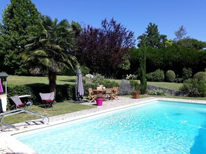 A swimming pool with a patio area, including a table and chairs, surrounded by lush greenery and trees. - La Maison Epellius (Collonges-au-Mont-d'Or)