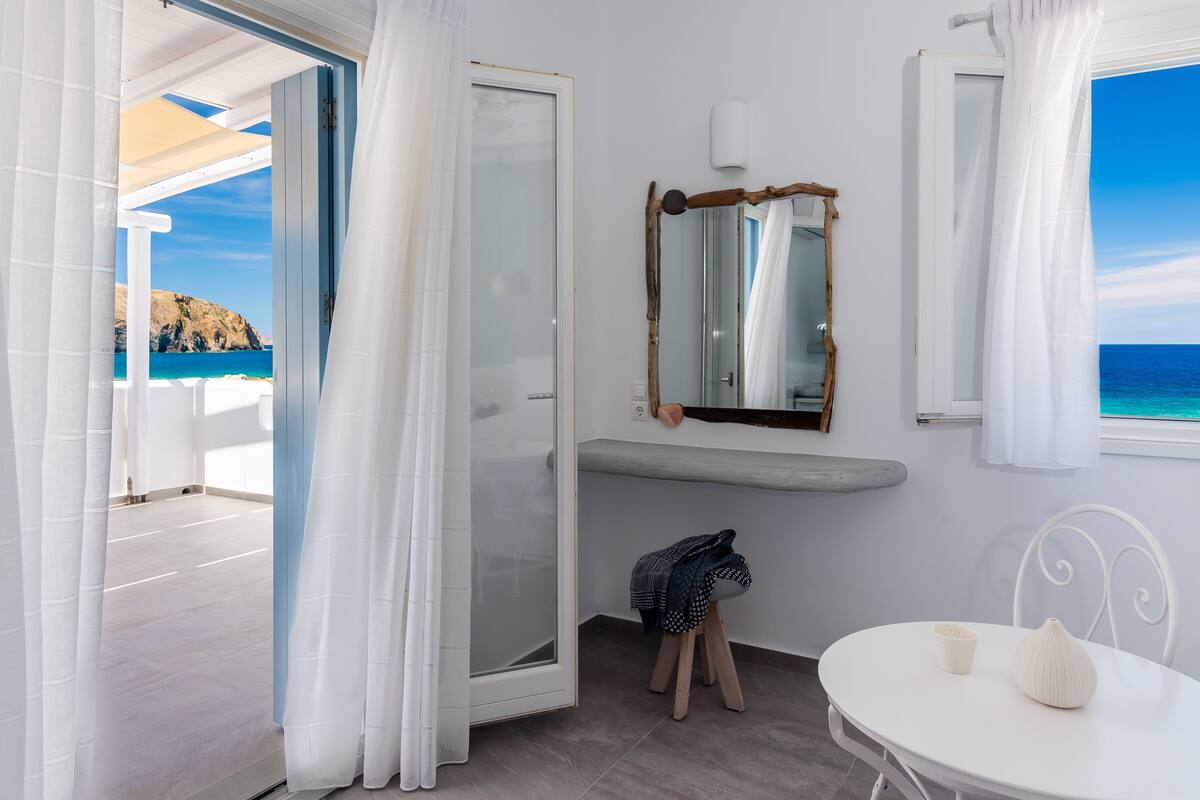 A room with a view of the sea, a white table with a vase, a mirror, and a small stool.