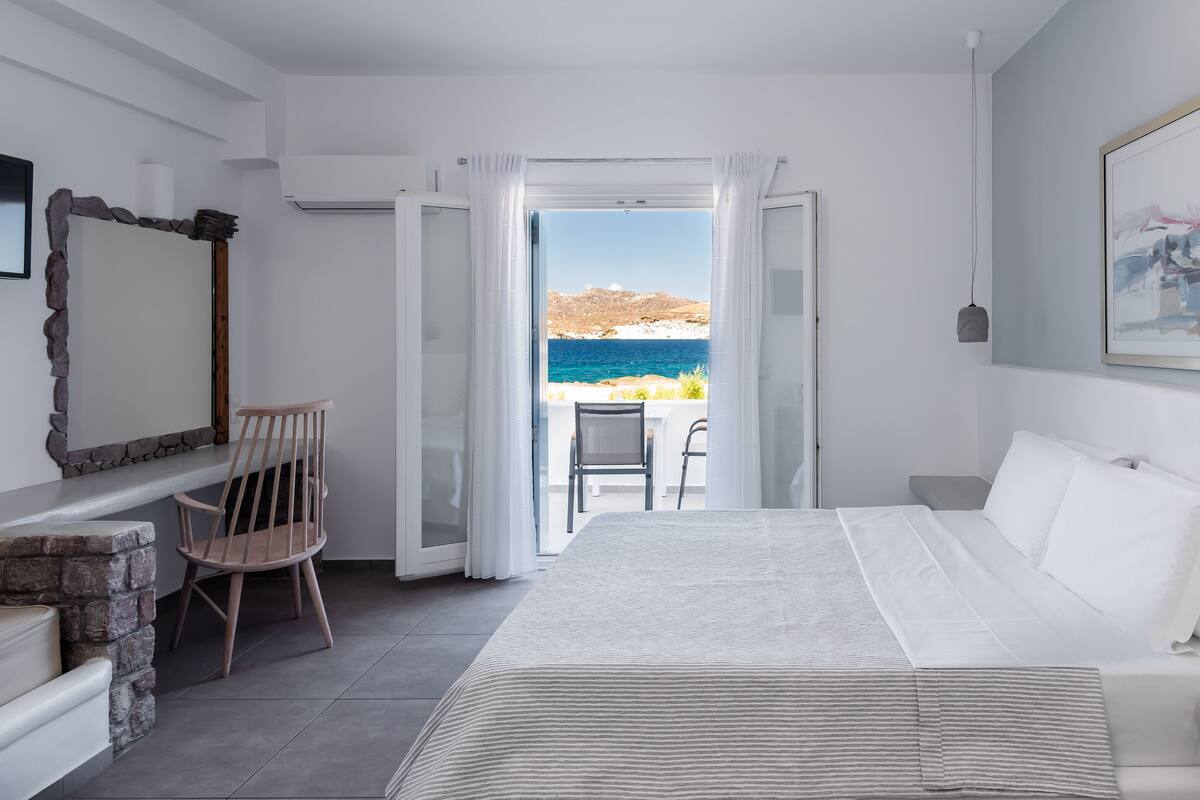 A bedroom with a bed, a chair, a small table, and a view of the sea.