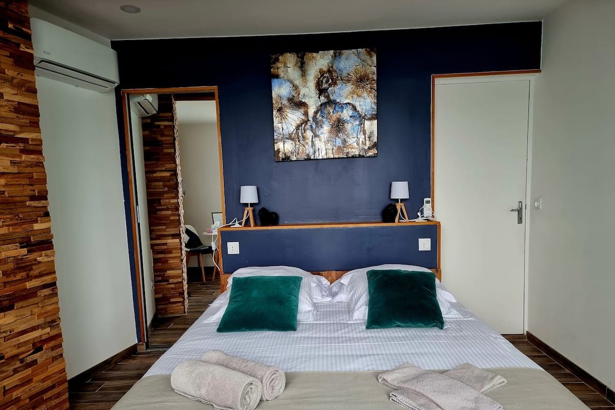 Habitación de hotel con una cama, dos almohadas verdes, un cuadro en la pared, un escritorio con una lámpara y una silla.