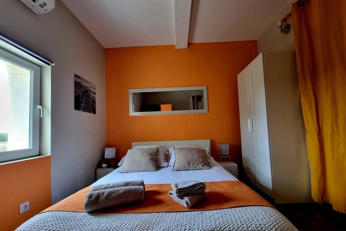 Un dormitorio con una pared naranja, una cama con ropa de cama blanca, un espejo sobre la cama y una ventana con cortinas.