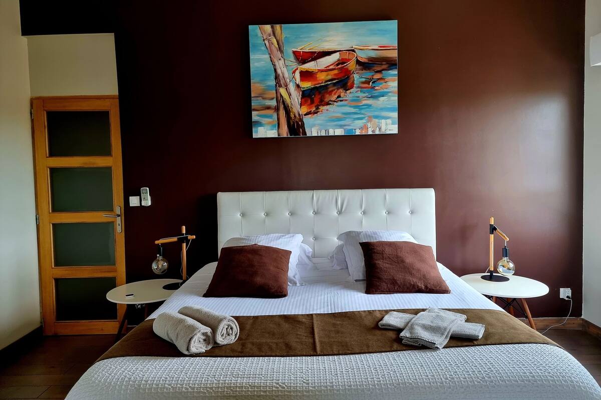 Habitación de hotel con una cama grande, dos almohadas, una mesita de noche, un cuadro en la pared y una puerta de madera.