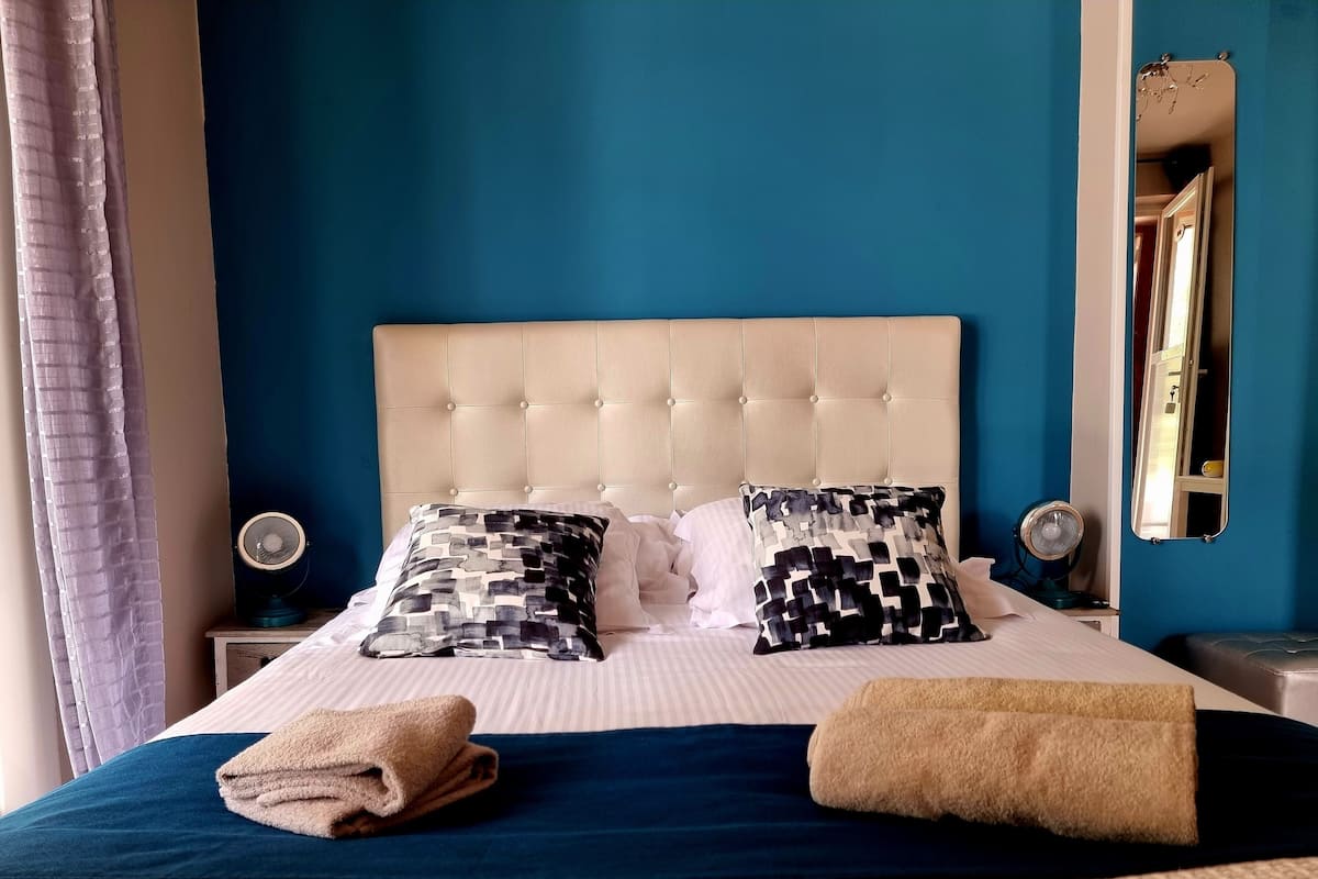 Una cama bien hecha con un cabecero acolchado, dos almohadas estampadas, una toalla doblada y una mesita de noche con un ventilador.
