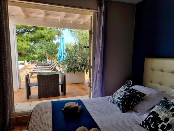Quarto com cama, almofadas e vista para um pátio externo com piscina, cadeiras e guarda-sóis.