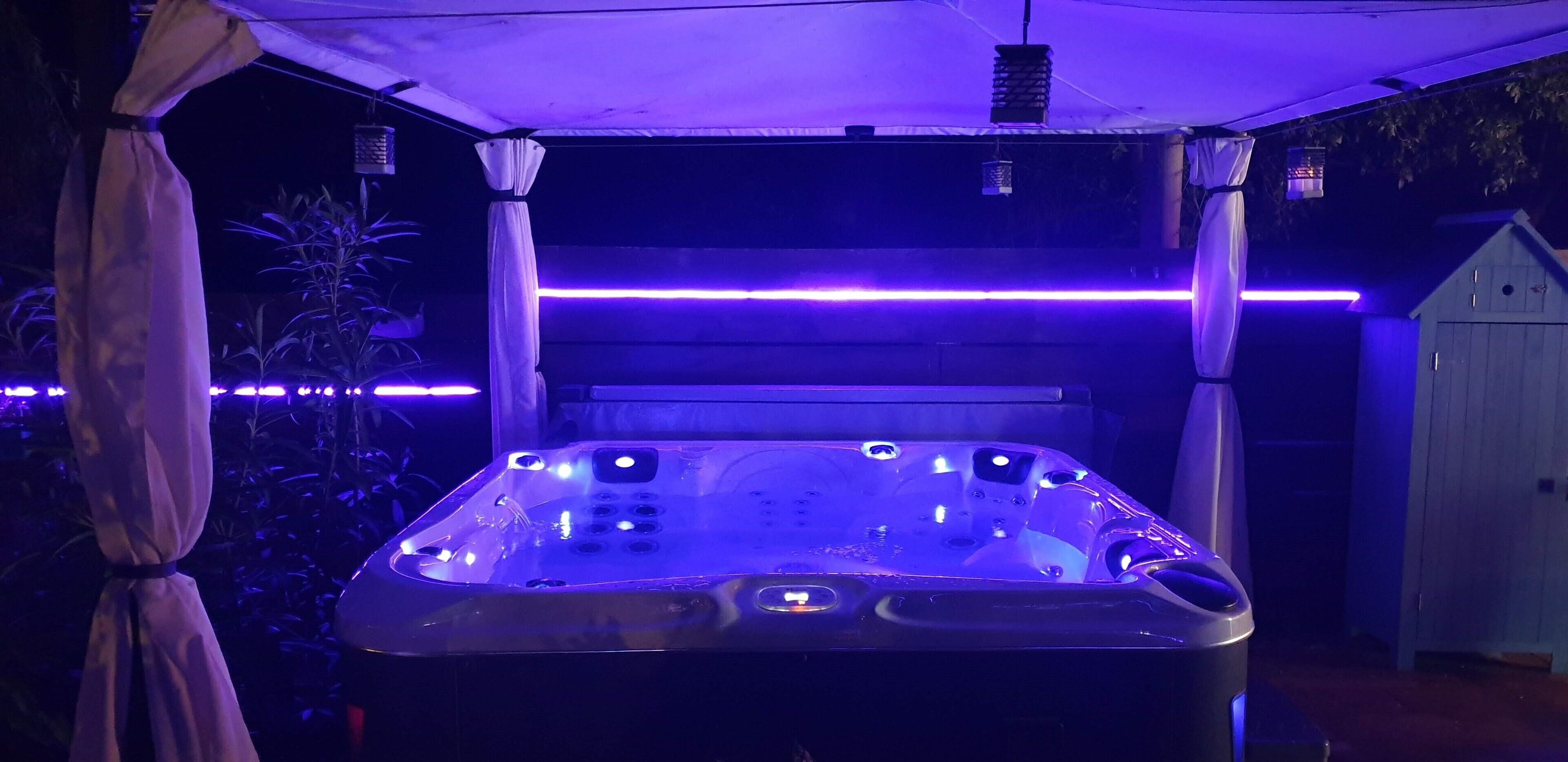 Ein Whirlpool unter einer Markise mit violetter Beleuchtung, umgeben von Pflanzen und einem Schuppen.