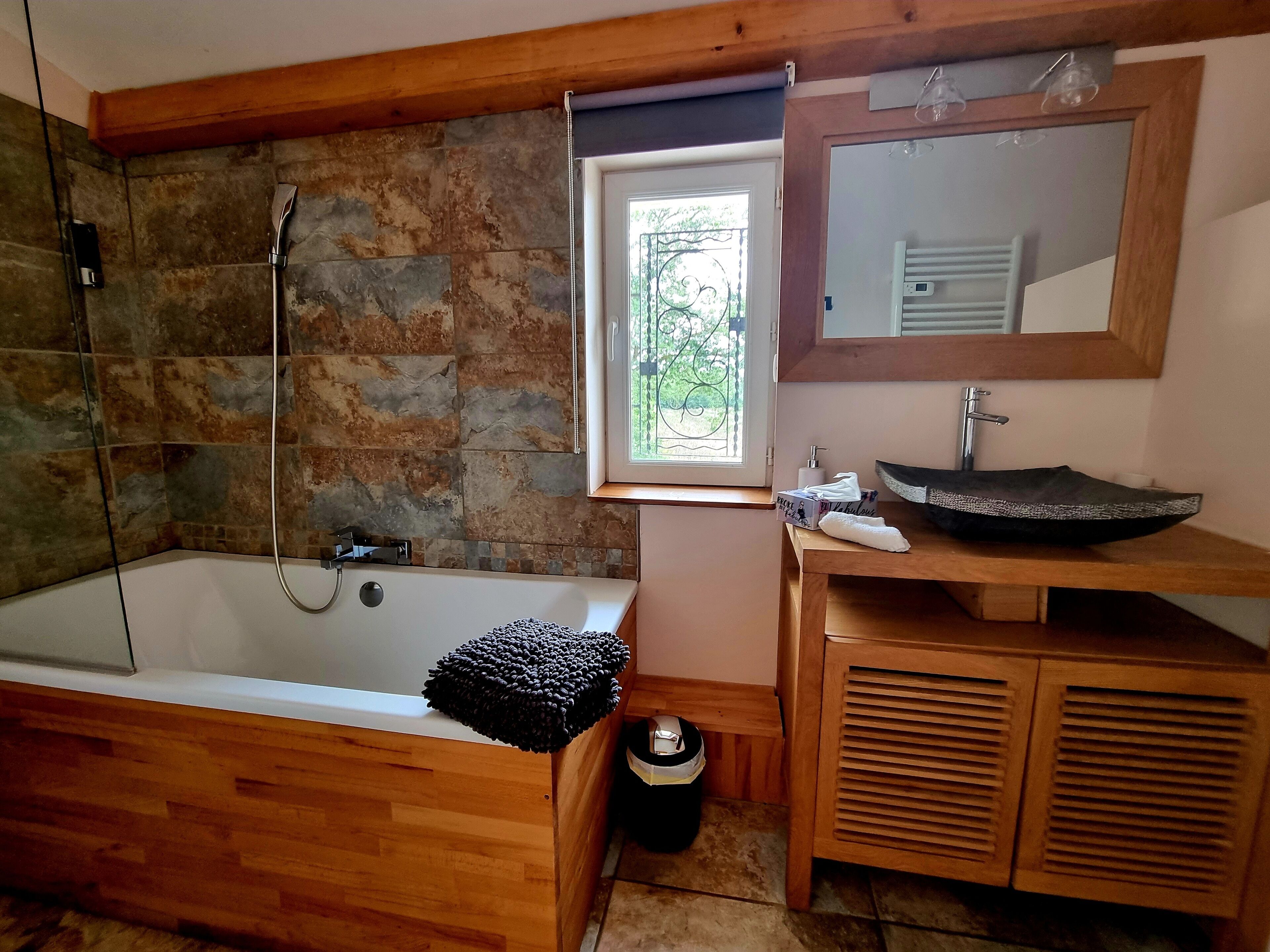 A bathroom with a stone wall, a bathtub, a wooden vanity, a sink, and a window with a view of greenery.