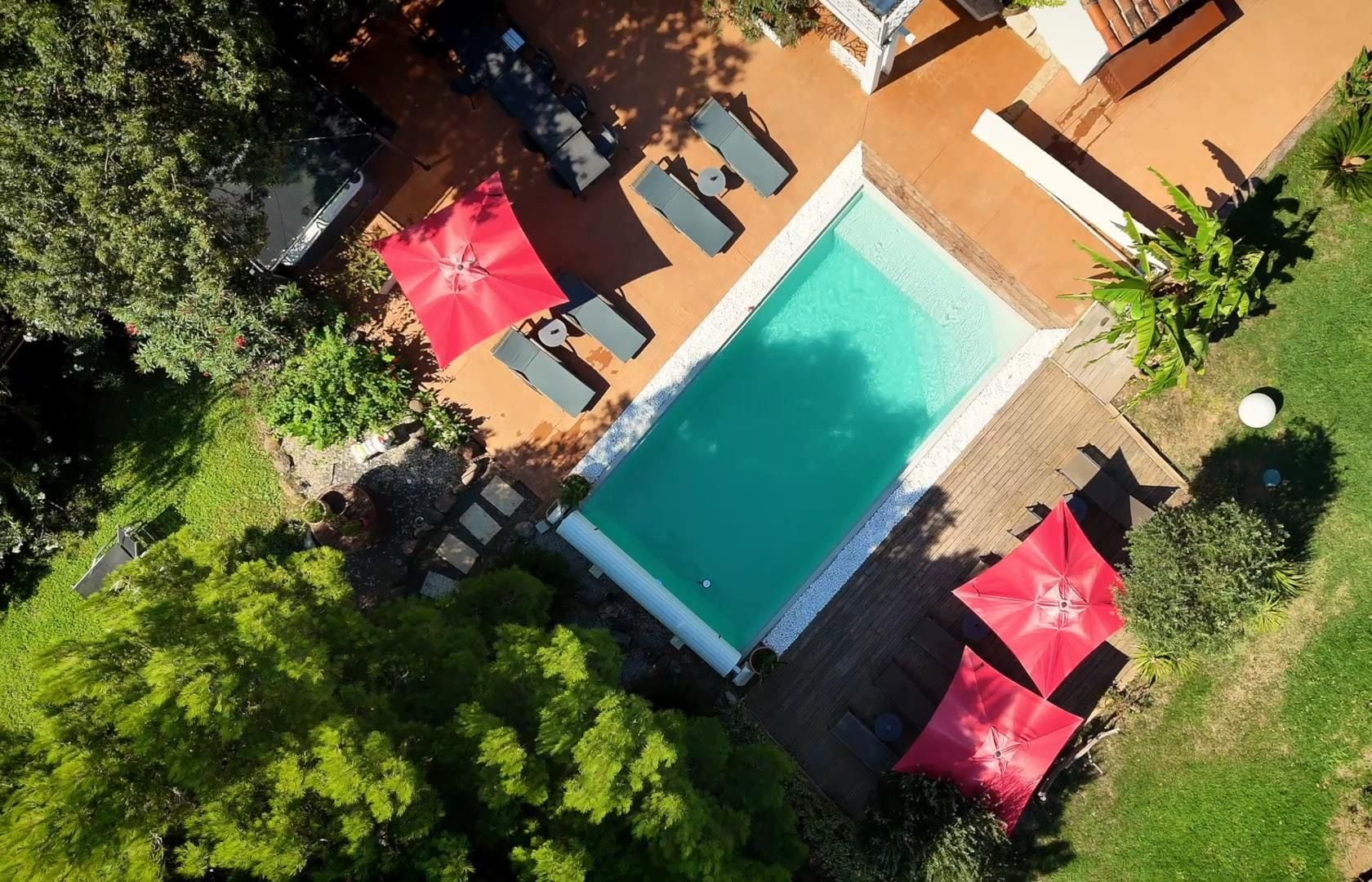 Vista aérea de un hotel con piscina rodeada de sillones de descanso y sombrillas rojas.