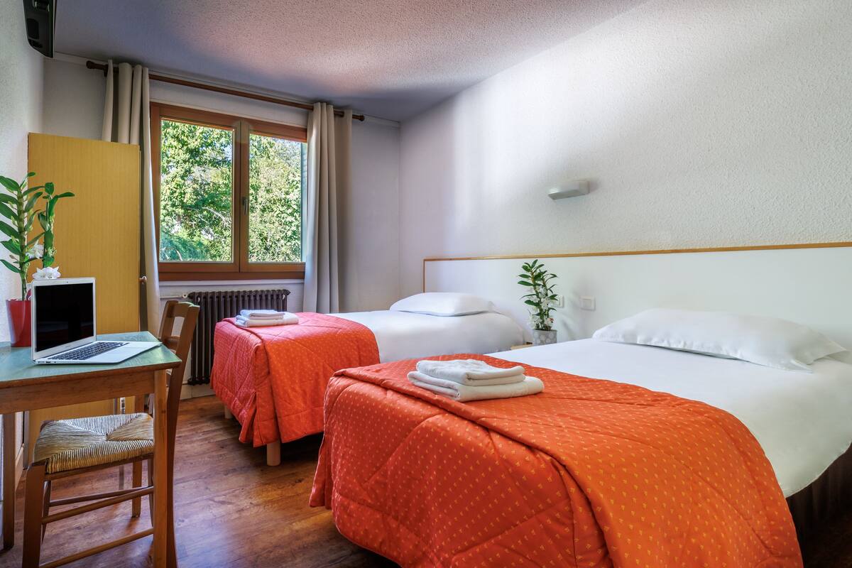 A hotel room with two beds, a desk with a laptop, a chair, and a window with curtains.