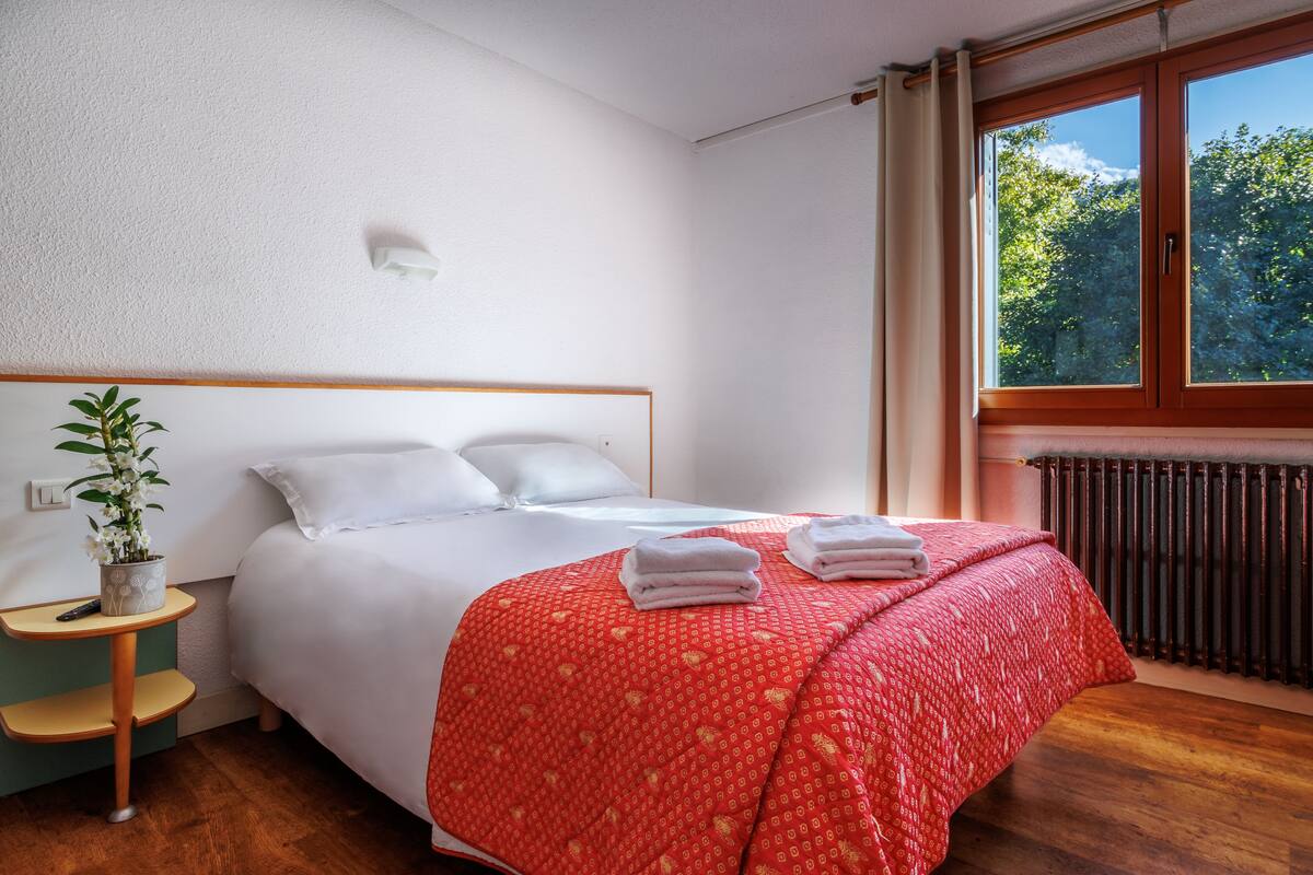 A bedroom with a bed, a bedside table with a plant, a window with a view of trees, and a radiator.