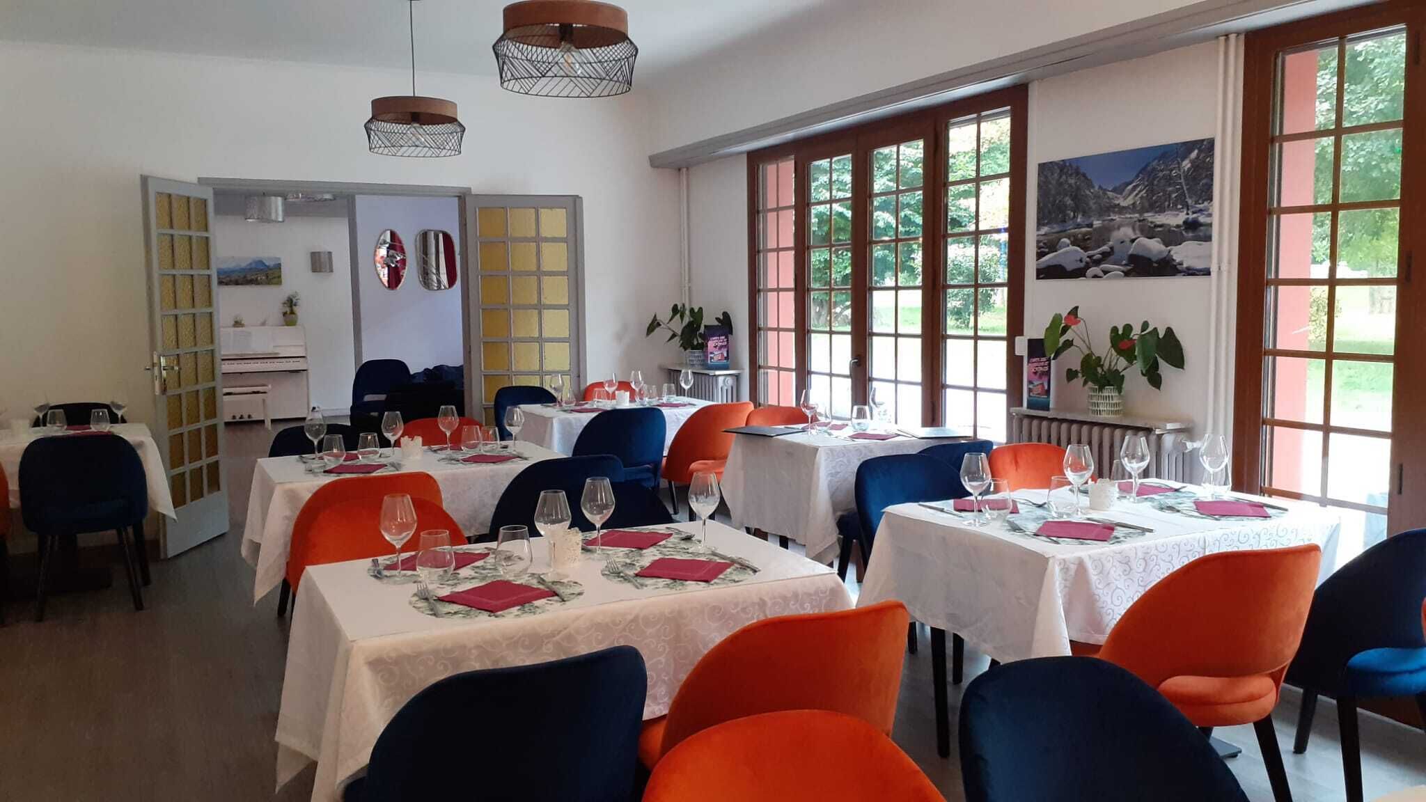 A dining area with tables set for multiple guests, featuring a mix of red and blue chairs, and large windows providing natural light.