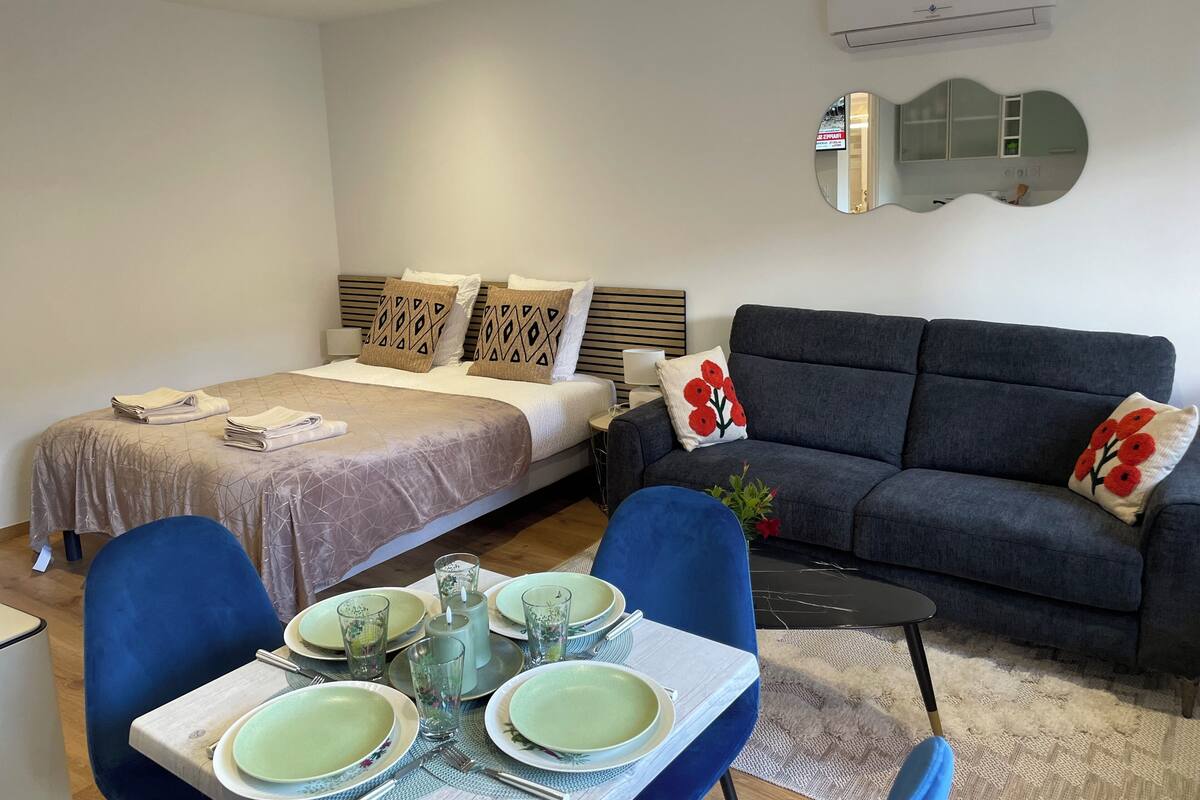 A modern bedroom with a bed, a grey sofa, a dining table set with plates and glasses, and a small side table.