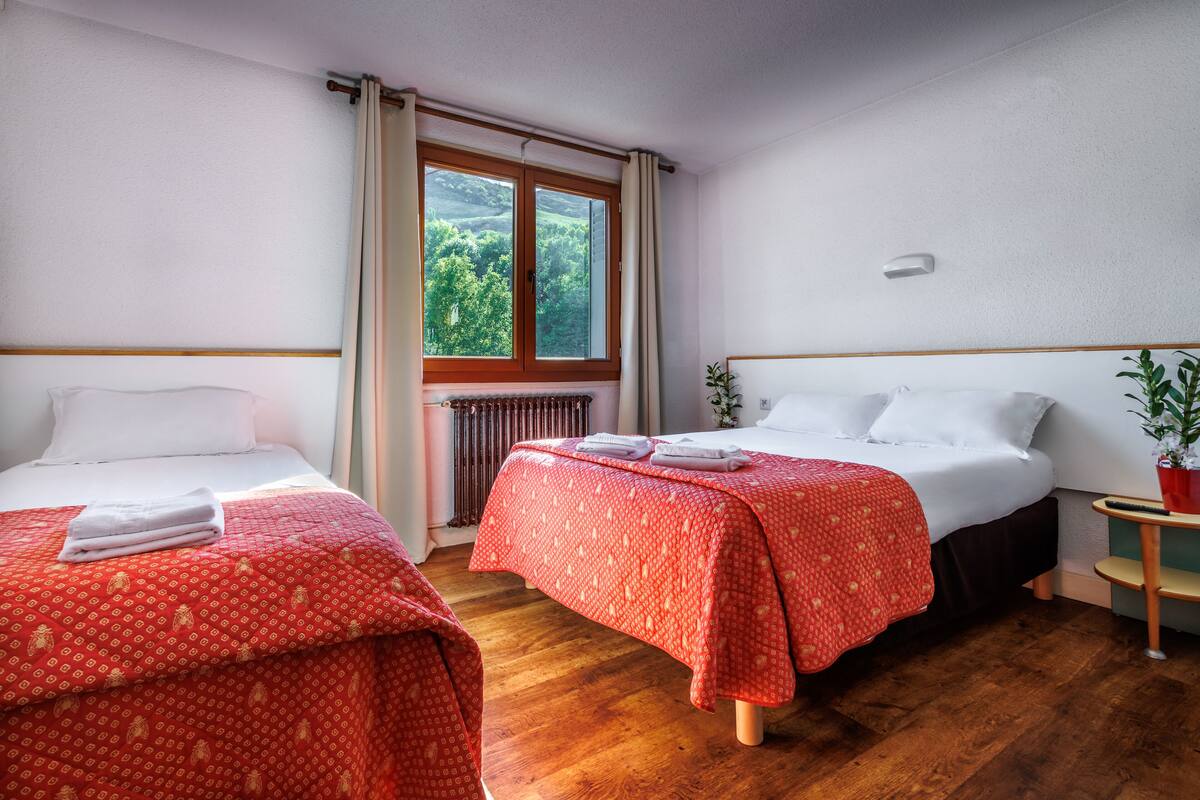 A hotel room with two beds, a window with curtains, a radiator, and a small table with a plant.