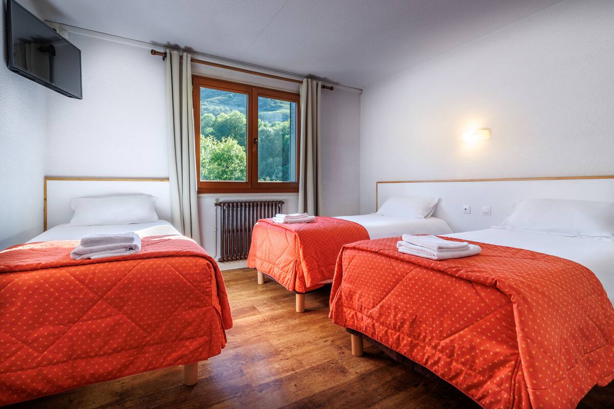 A hotel room with three beds, each with a red quilted bedspread and white pillows. There is a window with curtains, a flat-screen TV mounted on the wall, and a radiator near the window.