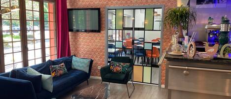 Habitació d'hotel moderna amb un sofà blau, butaques verdes, una taula de cafè de vidre i un televisor fixat a la paret.