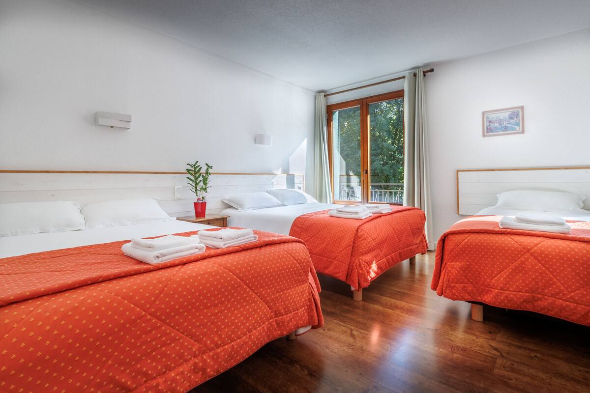 A hotel room with three beds, each with a red bedspread and white pillows. There is a window with curtains, a small plant, and a picture on the wall.