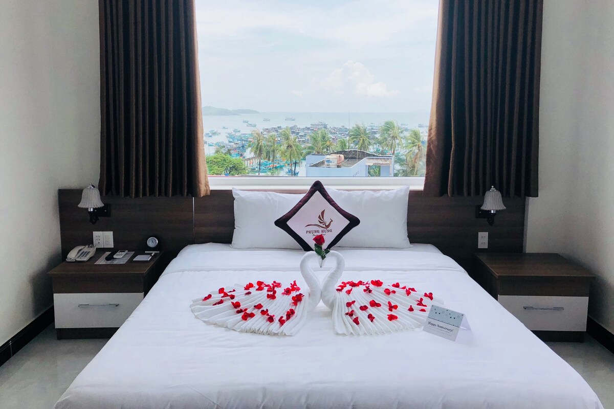 A hotel room with a bed, a nightstand, a lamp, a view of the sea, and a window with curtains.